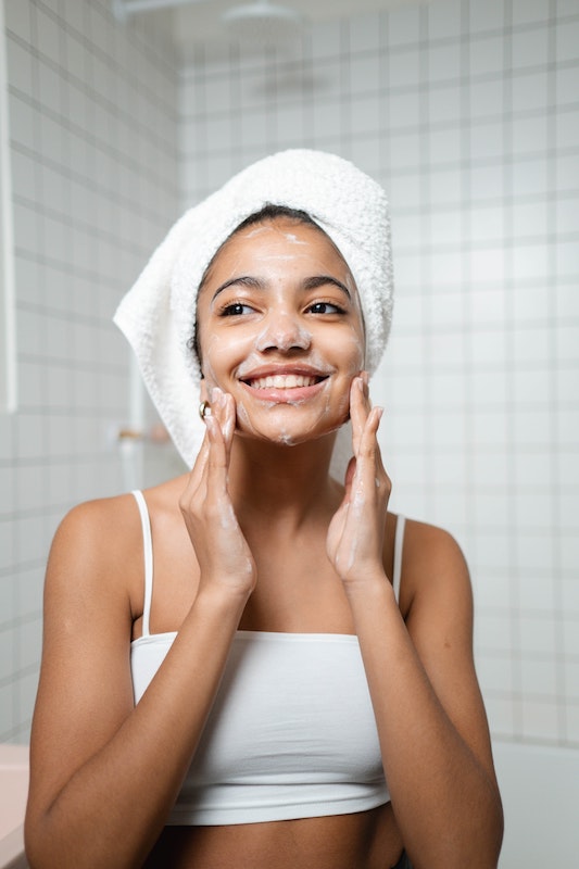 Girl with a white bath towel wrapped around her hair rubbing soap into her face and cheeks.